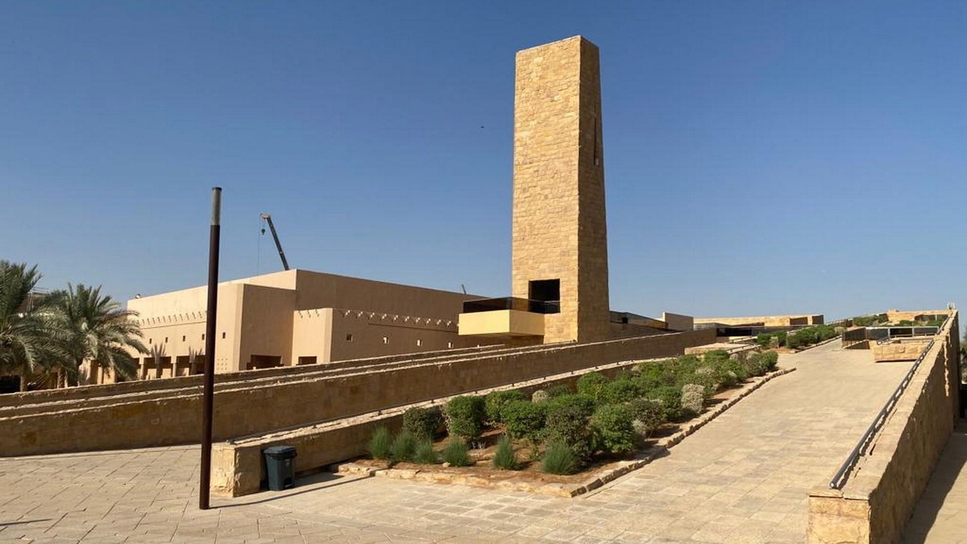 The Ministry of Culture headquarters in Riyadh: a tall stone-clad tower rising above the terraced plaza in front of the building, with palm trees to the left and paved ground in the foreground.